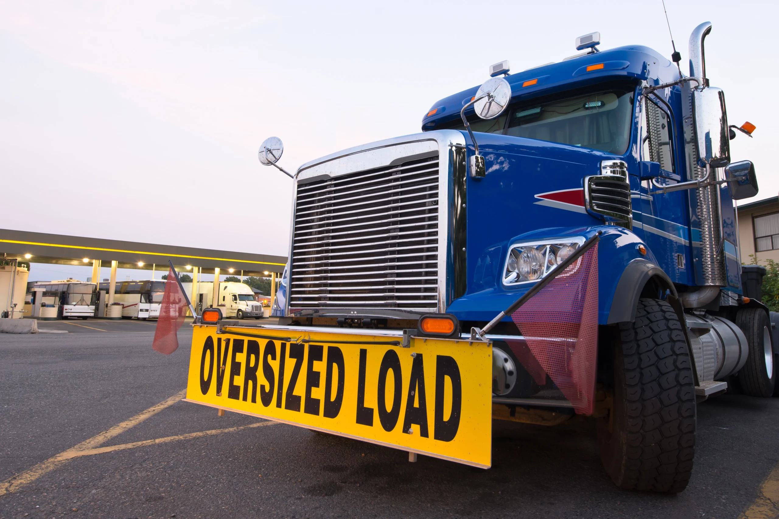 Blue semi-truck with oversized load sign parked at gas station, early evening light.