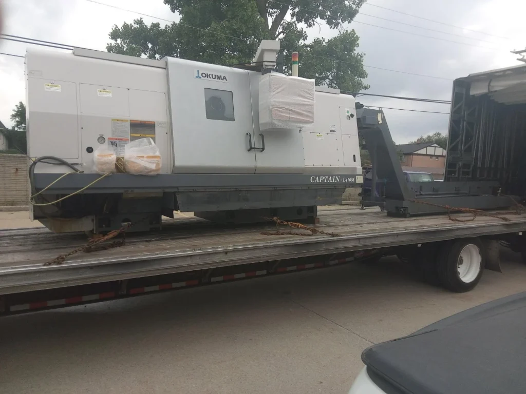 Heavy-duty Okuma CNC machine on flatbed truck for transport, secured with chains, outdoors on a cloudy day.