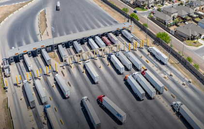 Aerial view of trucks lined up at a highway toll booth near a suburban neighborhood.
