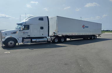 White semi-truck with enclosed trailer parked on sunny day at a rest area, blue skies and scattered clouds above.