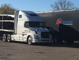 White semi-truck parked next to a black trailer with NAS logo in a sunny parking lot