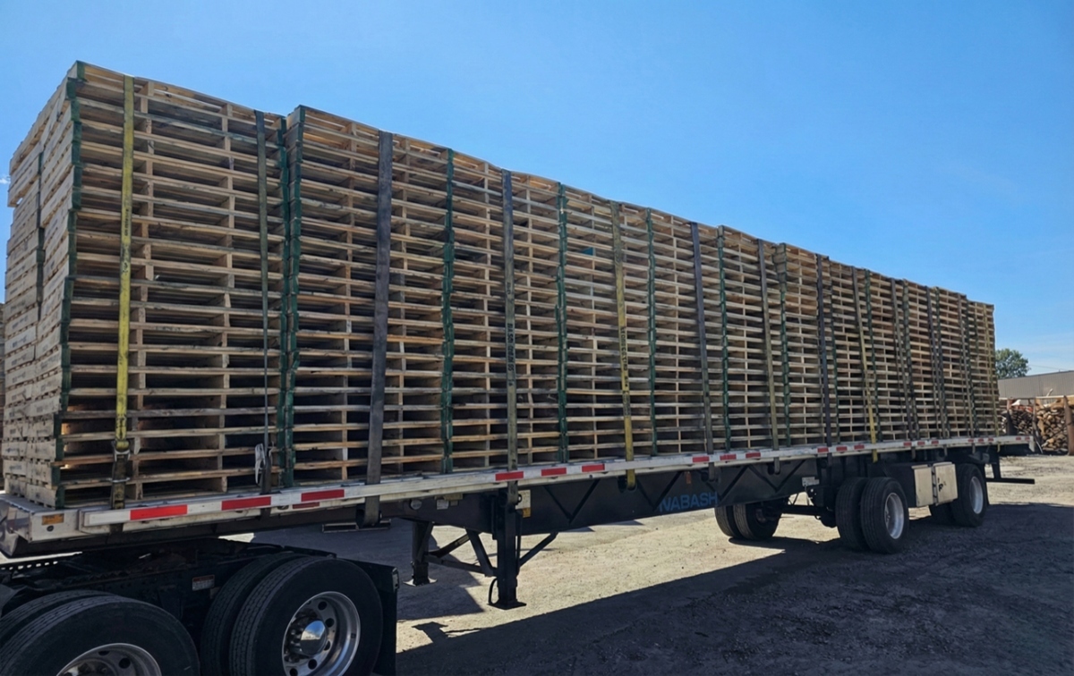 Flatbed trailer loaded with stacked wooden pallets under a clear blue sky