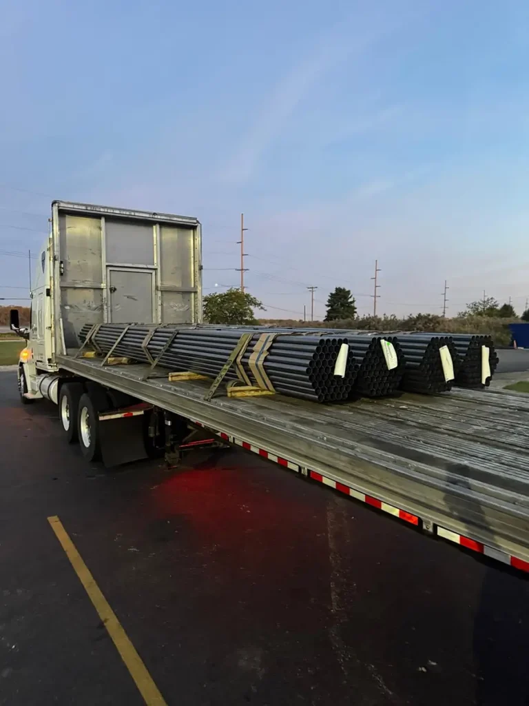 Flatbed truck loaded with stacked steel pipes for transportation at dusk.